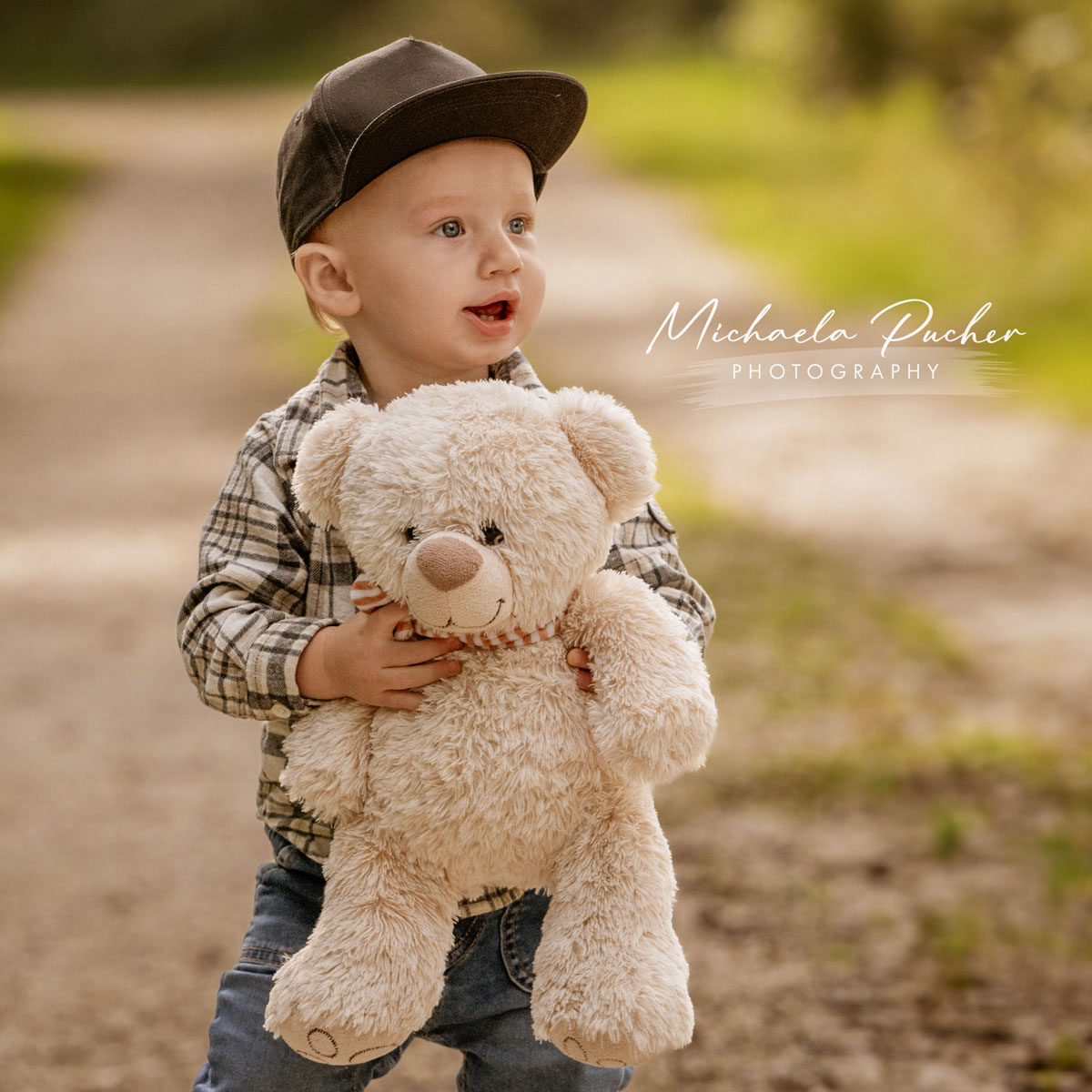 Kleiner Junge mit Kappe hält einen großen, flauschigen Teddybären auf einem sonnigen Waldweg und blickt neugierig zur Seite.